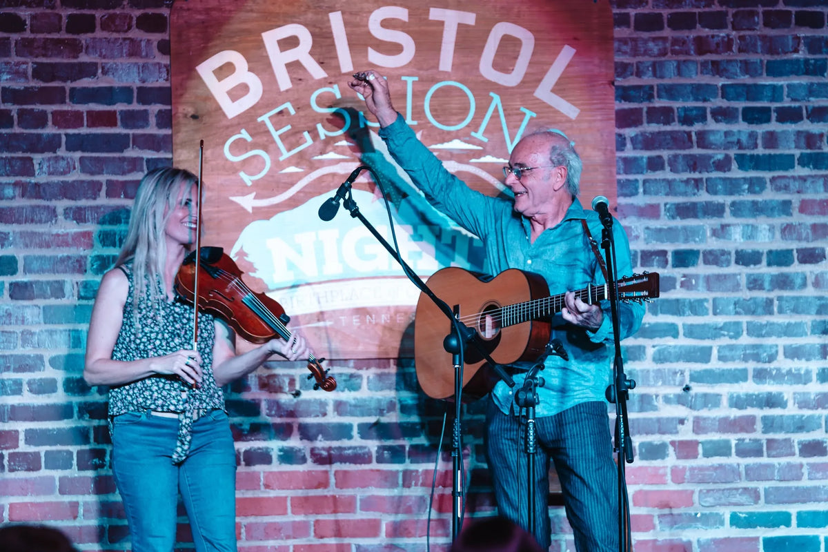Ed Snodderly raises a hand while playing guitar beside a fiddler under the Bristol Sessions Nights sign at Bristol Sessions Nights: The First Take, Bristol Listening Room, L.C. King Building, Bristol, Tennessee