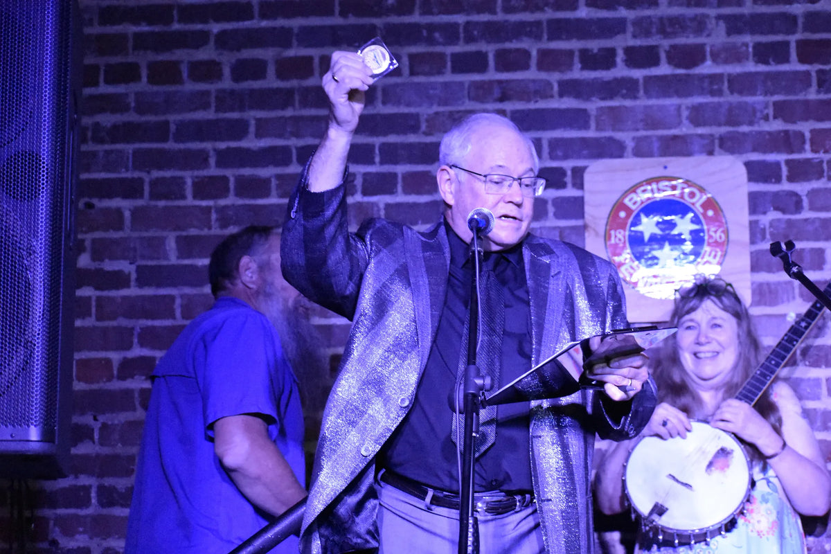 A storyteller holds up a keepsake during a lively moment on stage as a banjo player laughs at Bristol Sessions Nights: The Carter Sessions, Bristol Listening Room, L.C. King Building, Bristol, Tennessee